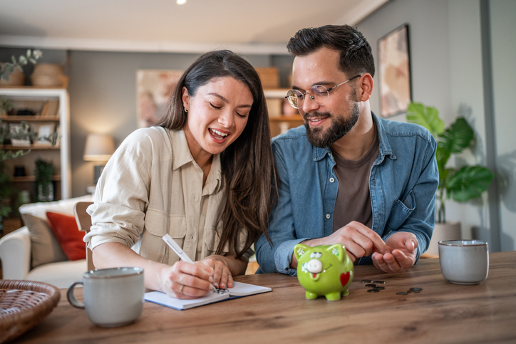 Young couple sitting at a wooden table, happily planning their home budget and saving money in a piggy bank, smiling and looking at each other