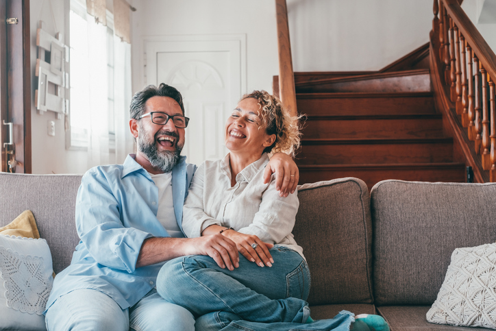 Happy caucasian couple laughing while sitting on sofa in the living room. Husband and wife laughing out loud sitting on couch at home. Man and woman spending leisure time at modern apartment Happy caucasian couple laughing while sitting on sofa in the living room. Husband and wife laughing out loud sitting on couch at home. Man and woman spending leisure time at modern apartment