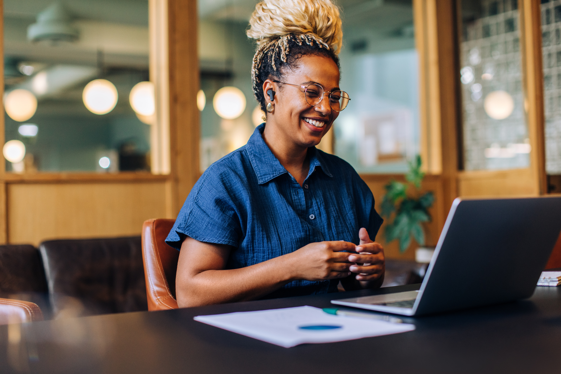 A cheerful woman engaging in a video conference in a contemporary office environment using a laptop.