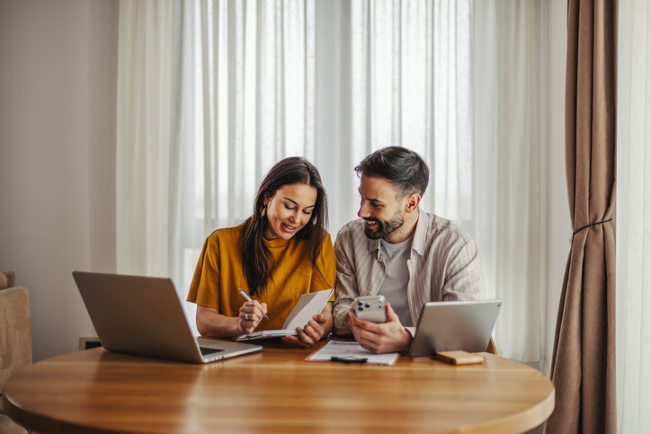 Young couple working together on their financial planning, reviewing documents and using digital devices like a laptop, smartphone, and tablet at home