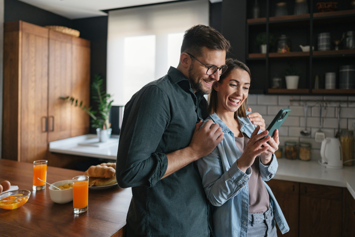 Young couple sharing a joyful moment while browsing their smartphone during breakfast in the kitchen, smiling and enjoying their time together over delicious food and drinks